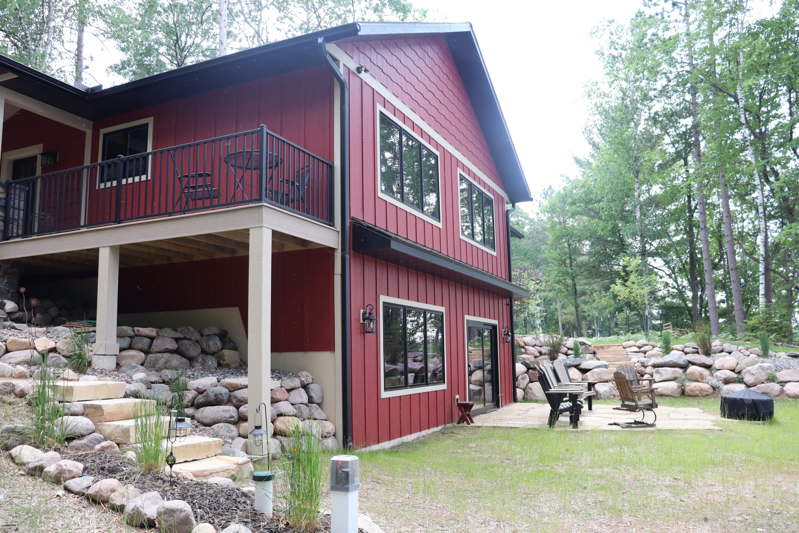 A red two-story house with large windows, custom tile showers, a covered balcony, and stone landscaping sits in a wooded area with a fire pit and outdoor seating nearby.