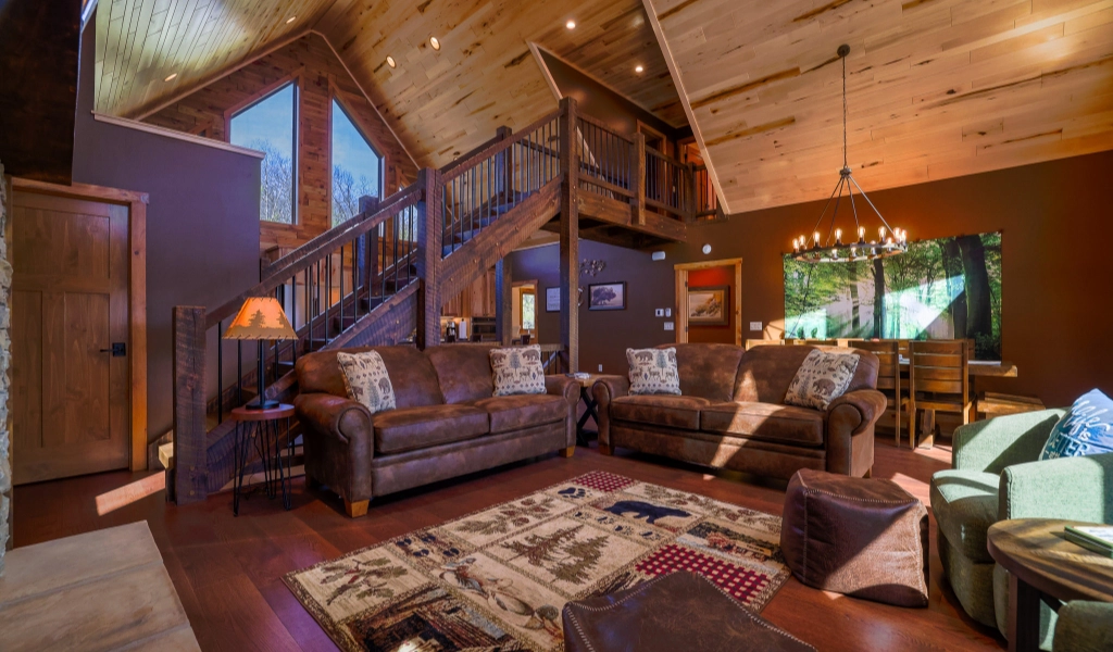 Rustic living room with wood-paneled walls and ceiling, two brown sofas, patterned rug, and a staircase leading to a loft area. Dining table, chandelier, and nearby custom tile showers add charm to the inviting space.