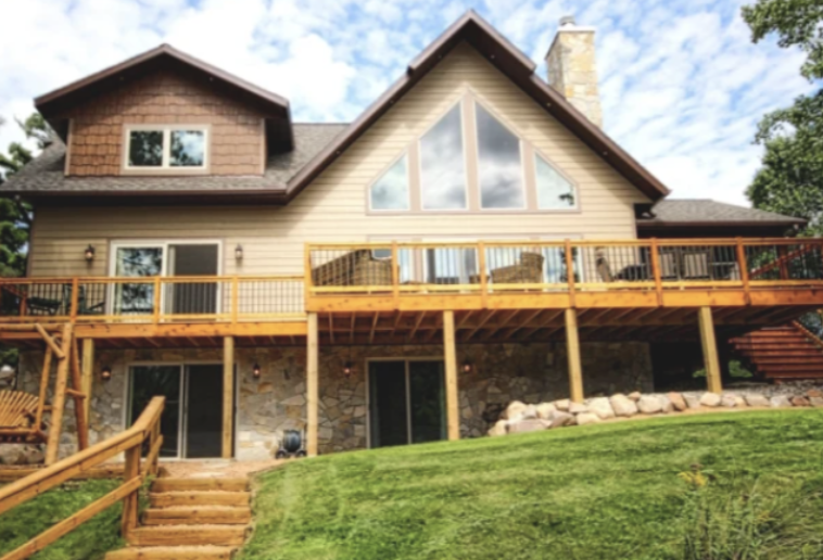 A two-story house with large triangular windows, a wooden deck, and stone foundation sits on a grassy slope surrounded by trees under a partly cloudy sky, featuring custom tile showers for added luxury inside.