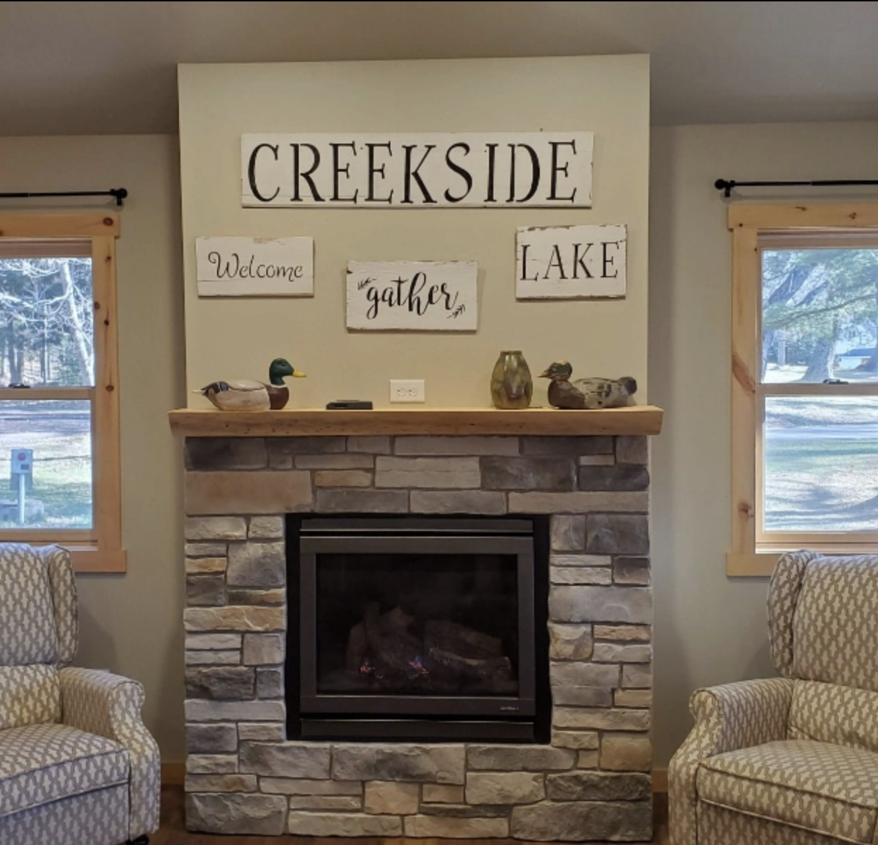 Stone fireplace with a wooden mantel, decorative ducks, and framed signs reading "Creekside," "Welcome," "gather," and "Lake" between two windows and armchairs—perfect to pair with baths featuring custom tile showers.
