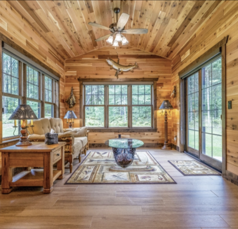 A cozy sunroom with wood-paneled walls and ceiling, large windows, a sofa set, glass coffee table, area rug, and sliding glass doors opening to a green outdoor view—perfect for relaxing after enjoying custom tile showers nearby.