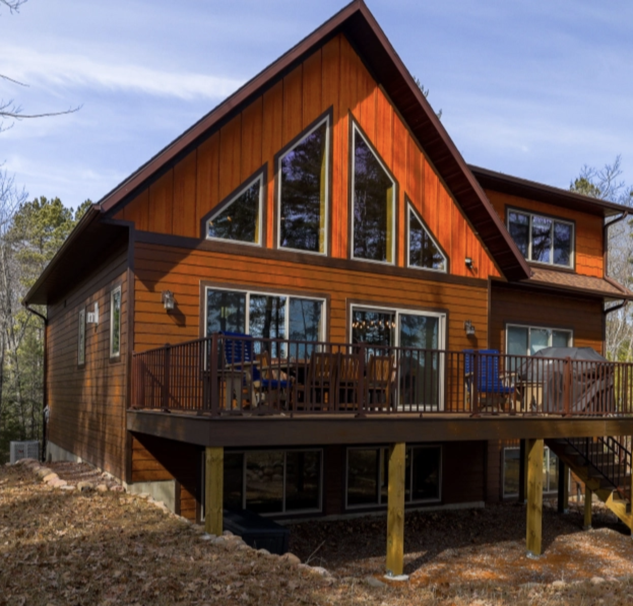 A modern two-story wooden house with large triangular windows, custom tile showers, and a spacious elevated deck with outdoor furniture, surrounded by trees.