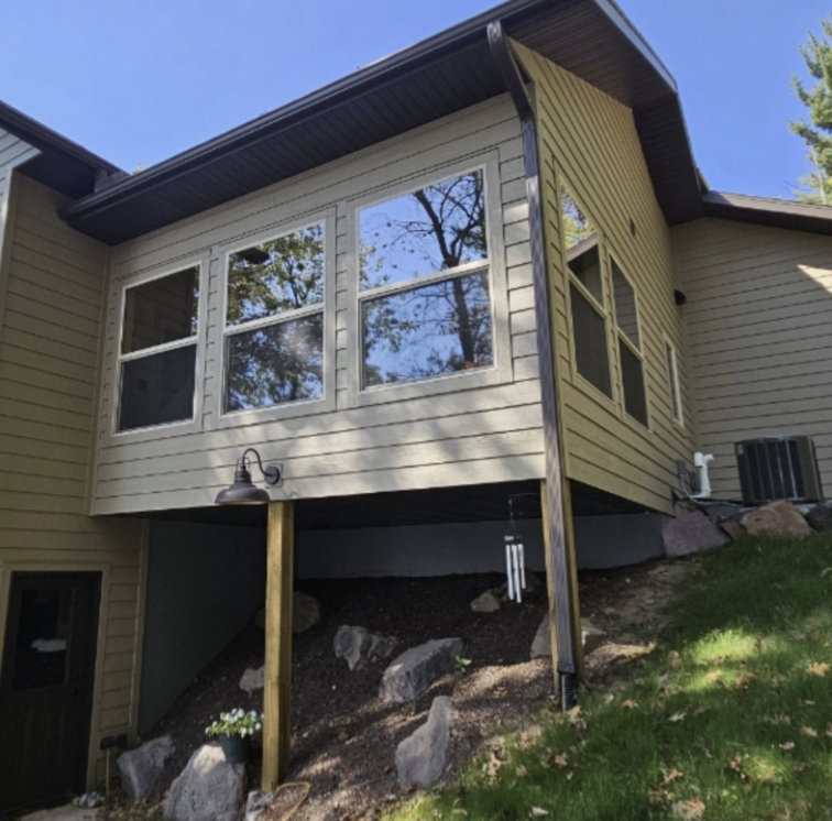 A house with a raised, enclosed sunroom featuring large windows, supported by two wooden posts, sits on a sloped yard with rocks and a wind chime underneath—inside, discover inviting spaces and custom tile showers.