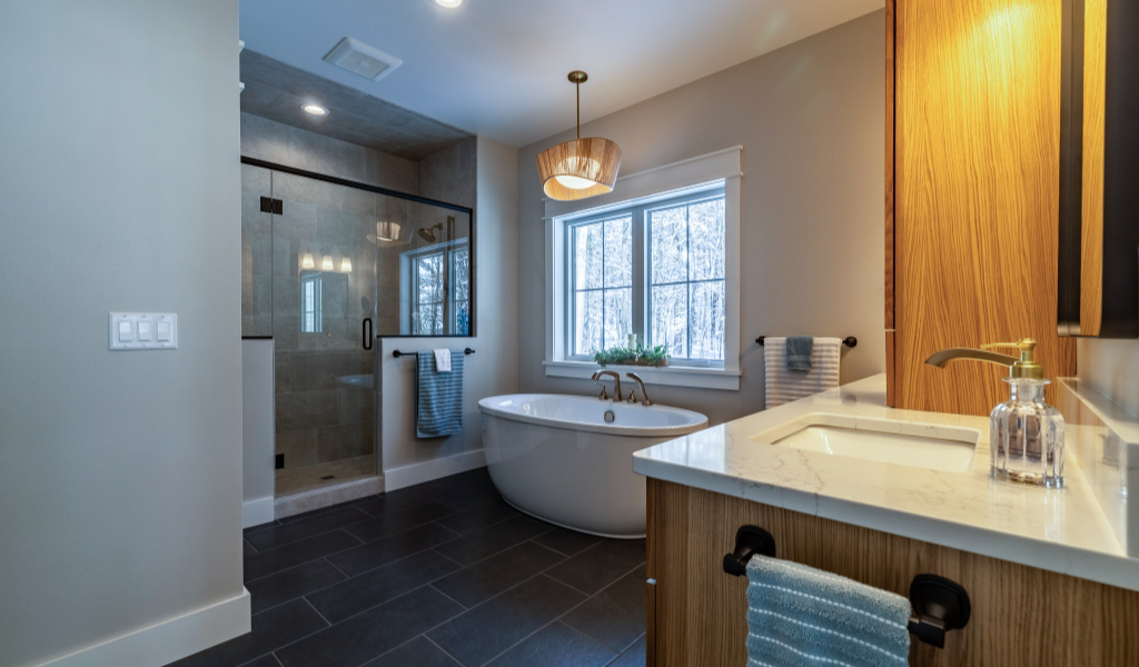 Modern bathroom with a freestanding tub by a window, custom tile shower enclosed in glass, wood cabinetry, and a marble countertop under warm lighting.