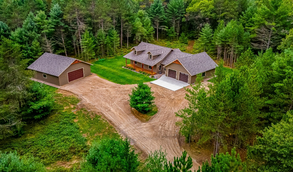 Aerial view of a house and detached garage surrounded by dense trees, with a dirt driveway, small lawn area, and potential for custom tile showers inside for added luxury.