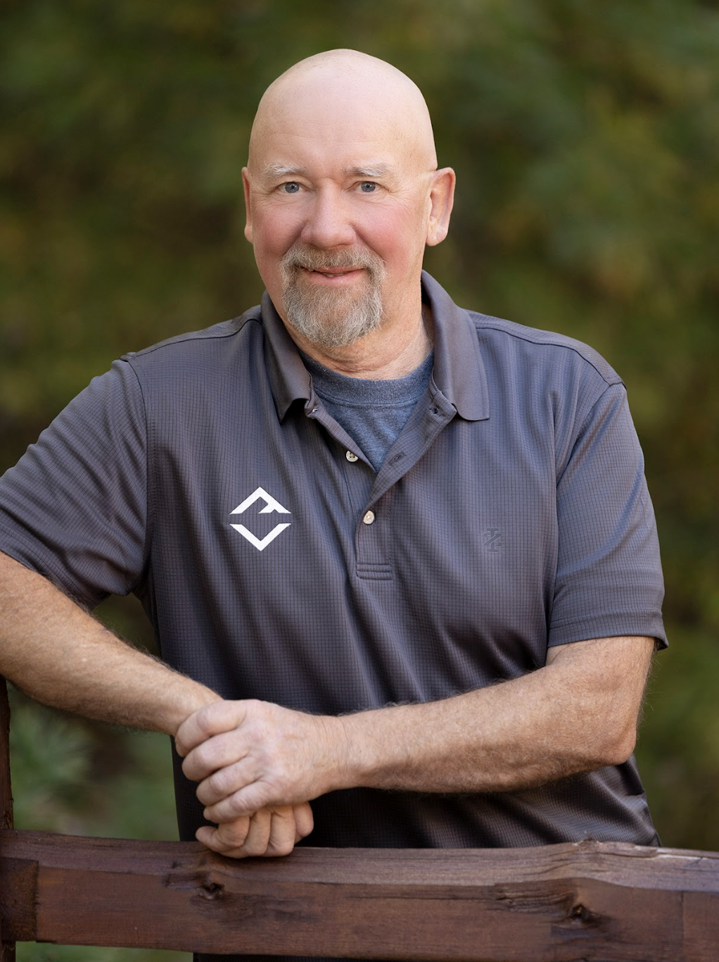 A bald man with a goatee, wearing a gray polo shirt, stands outdoors with greenery in the background, resting his arms on a wooden fence—perhaps contemplating ideas for custom tile showers.