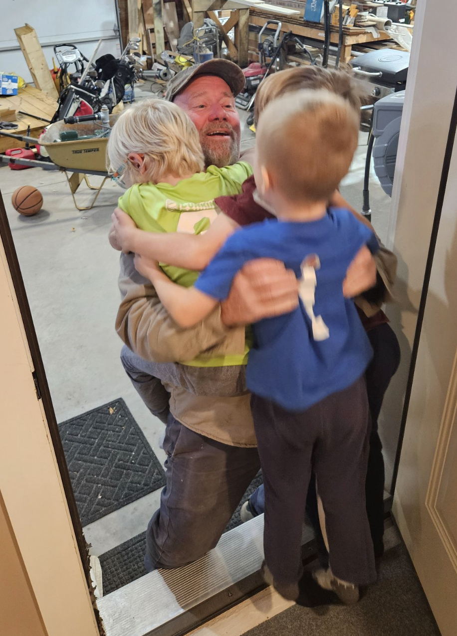 An adult kneels in a garage with a smile, hugging three young children who are enthusiastically greeting him at the doorway, just steps away from their home’s custom tile showers.