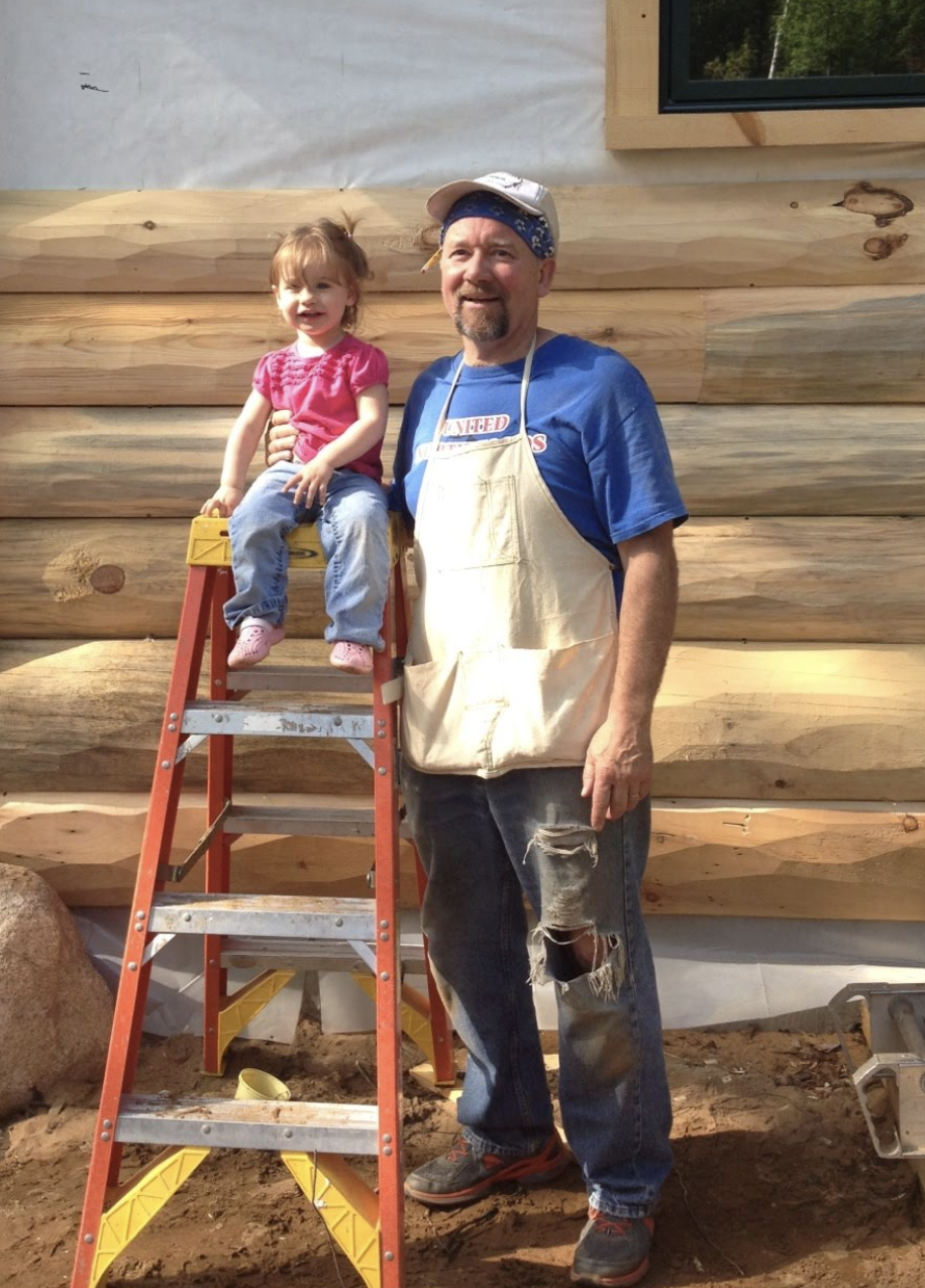 A man in an apron stands next to a young girl sitting on a step ladder in front of a log cabin under construction, discussing plans for custom tile showers inside the new home.