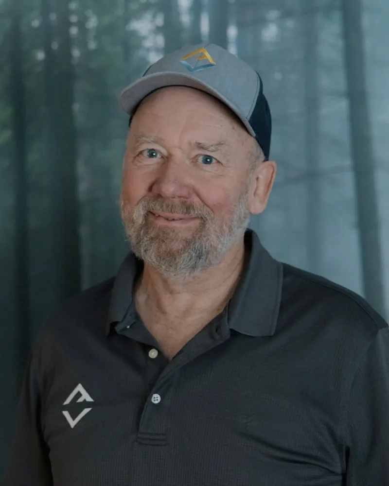 An older man with a gray beard wears a gray logo shirt and cap, standing in front of a blurred forest background—ready to discuss custom tile showers.